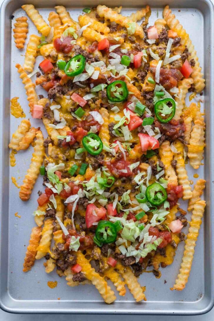Crinkle-cut fries on a baking sheet topped with melted cheese, ground beef, diced tomatoes, shredded lettuce, salsa, and sliced jalapeños.