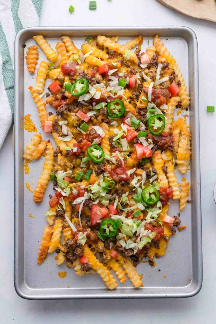 A metal baking sheet with crinkle-cut fries topped with melted cheese, ground beef, diced tomatoes, shredded lettuce, sliced jalapeños, and green onions, creating loaded nacho fries.