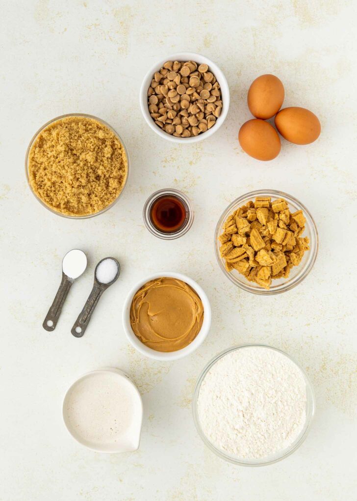 Nine bowls and cups of baking ingredients, including brown sugar, peanut butter chips, eggs, vanilla, cubed cookie pieces, peanut butter, flour, milk, plus two measuring spoons with salt and baking soda, arranged on a white surface.