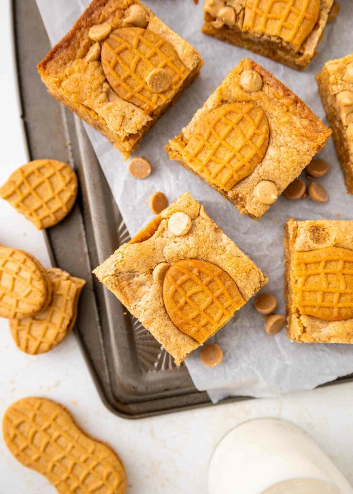 Cookie bars topped with peanut-shaped cookies and peanut butter chips on parchment paper, with more cookies and a glass of milk nearby.