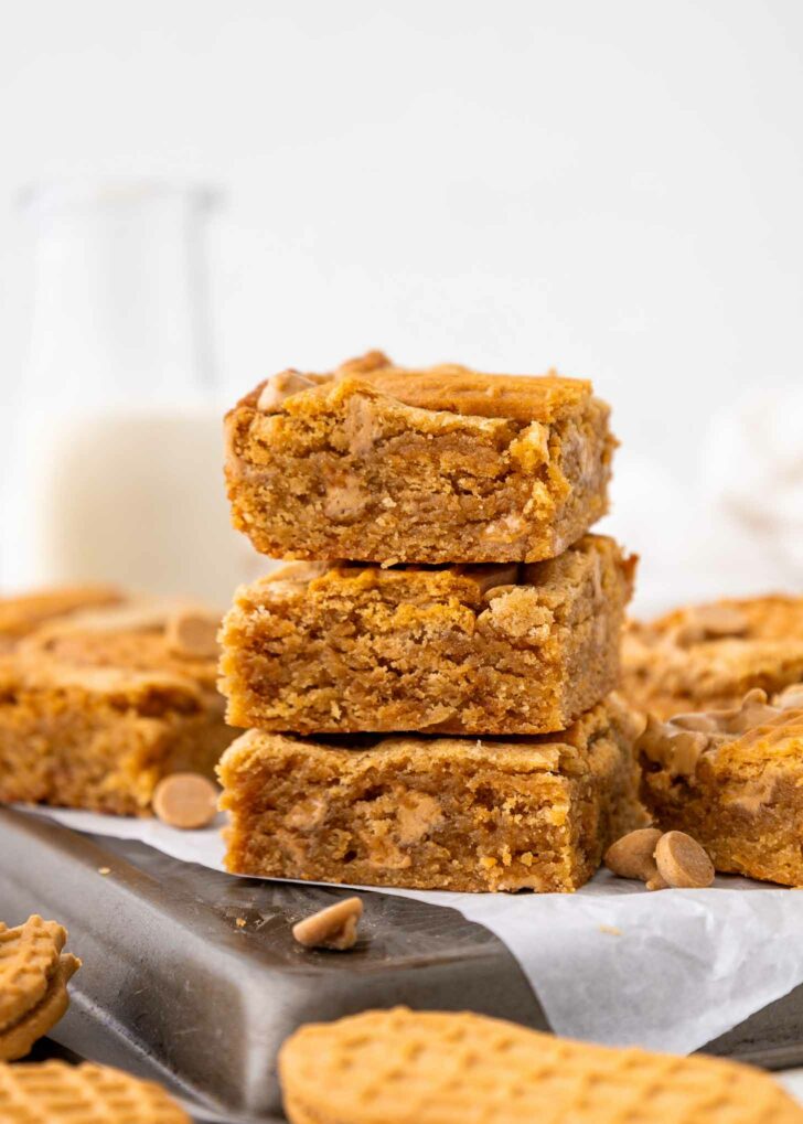 Three thick cookie bars are stacked on a baking sheet lined with parchment paper, surrounded by more blondies, peanut butter chips, and a few round cookies. A glass bottle of milk is blurred in the background.