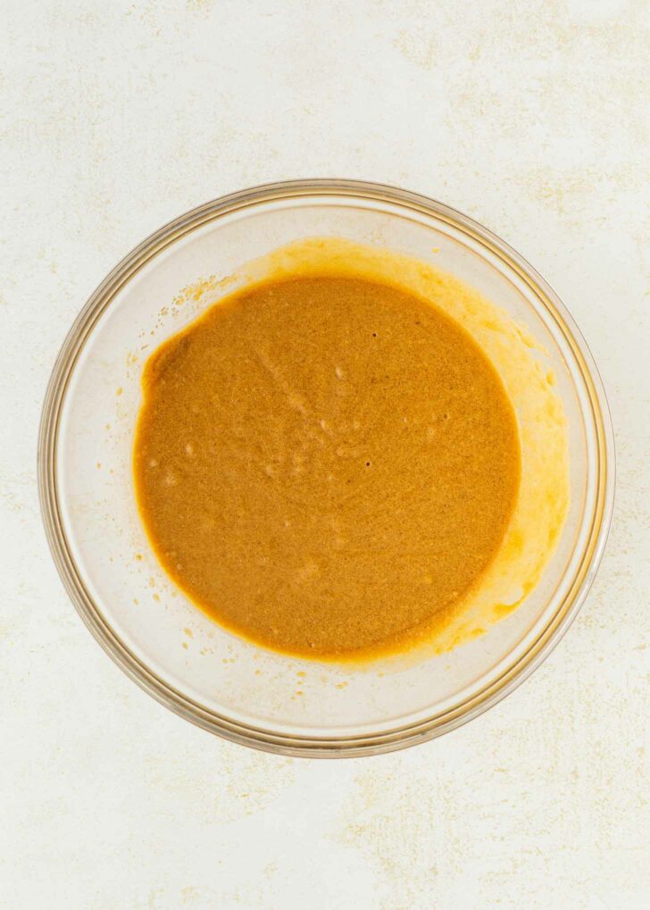 A clear glass bowl filled with a smooth, light brown batter sits on a white textured surface.
