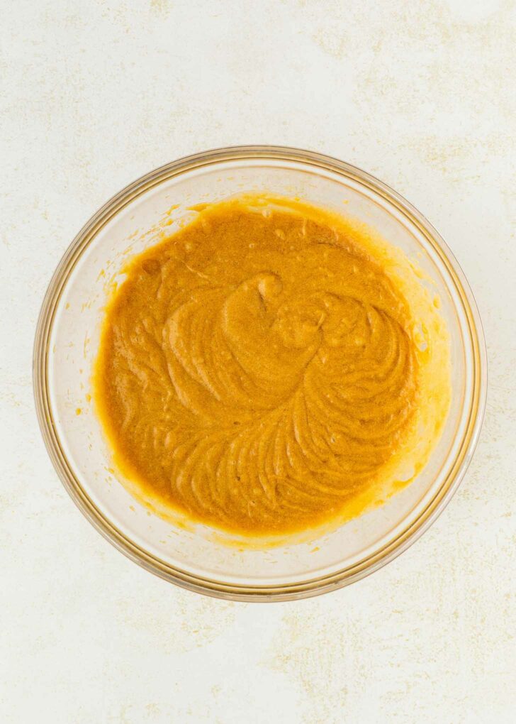 A glass mixing bowl filled with smooth, light brown batter sits on a white surface. The batter appears to be freshly mixed with visible swirls and a creamy texture.