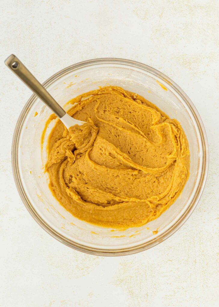 A glass bowl filled with smooth, orange-brown cookie dough sits on a light-colored surface. A white spatula with a metal handle rests in the dough.