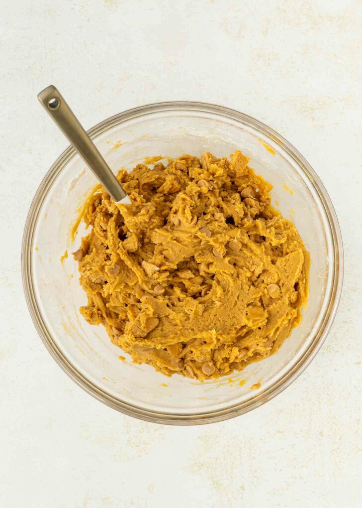 A glass mixing bowl filled with thick, golden-brown cookie dough and a metal spoon, set on a light-colored surface.