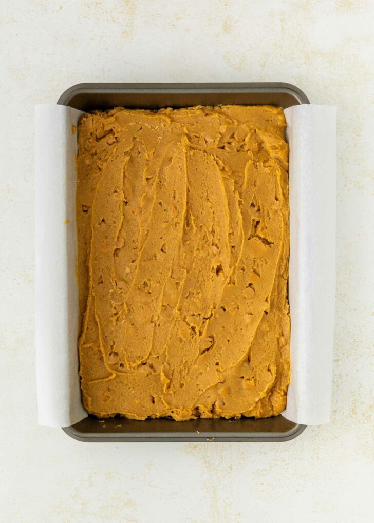 A rectangular metal baking pan lined with parchment paper and filled with unbaked, spread-out peanut butter cookie mixture on a light-colored surface.