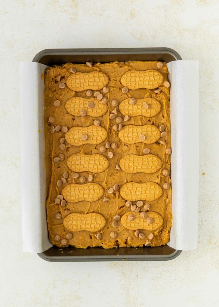 A rectangular baking pan lined with parchment paper holds unbaked dough topped with whole peanut-shaped cookies and chocolate chips, arranged neatly on a light-colored surface.