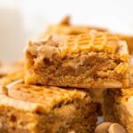 A close-up of golden brown peanut butter bars stacked on top of each other, with a bite taken from the top piece. Peanut butter chips are scattered around the blondies.