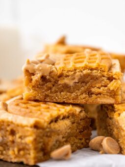 A close-up of golden brown peanut butter bars stacked on top of each other, with a bite taken from the top piece. Peanut butter chips are scattered around the blondies.