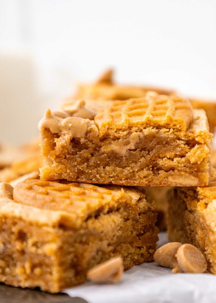 A close-up of golden brown peanut butter bars stacked on top of each other, with a bite taken from the top piece. Peanut butter chips are scattered around the blondies.