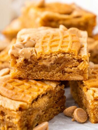 A close-up of stacked peanut butter bars with a peanut-shaped cookie pressed on top of each bar, surrounded by peanut butter chips.