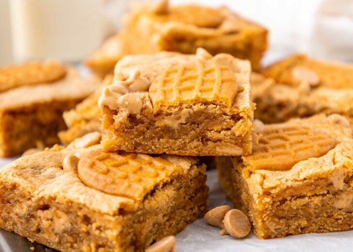 A close-up of stacked peanut butter bars with a peanut-shaped cookie pressed on top of each bar, surrounded by peanut butter chips.