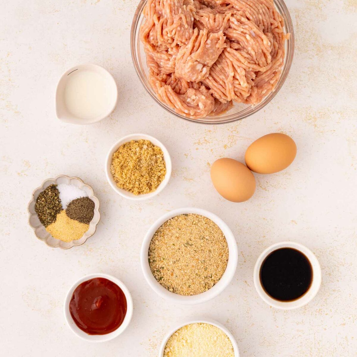 Top-down view of various meatloaf ingredients on a light surface, including raw ground meat in a bowl, two eggs, seasonings, breadcrumbs, milk, ketchup, Worcestershire sauce, and grated cheese in small dishes.