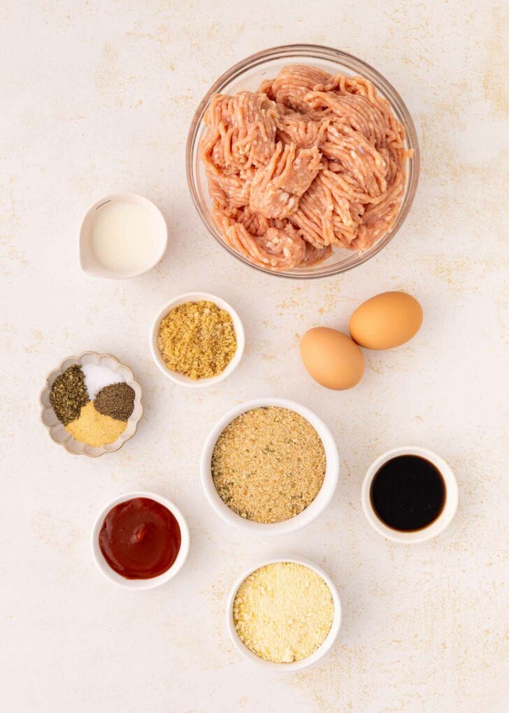 Top-down view of various meatloaf ingredients on a light surface, including raw ground meat in a bowl, two eggs, seasonings, breadcrumbs, milk, ketchup, Worcestershire sauce, and grated cheese in small dishes.