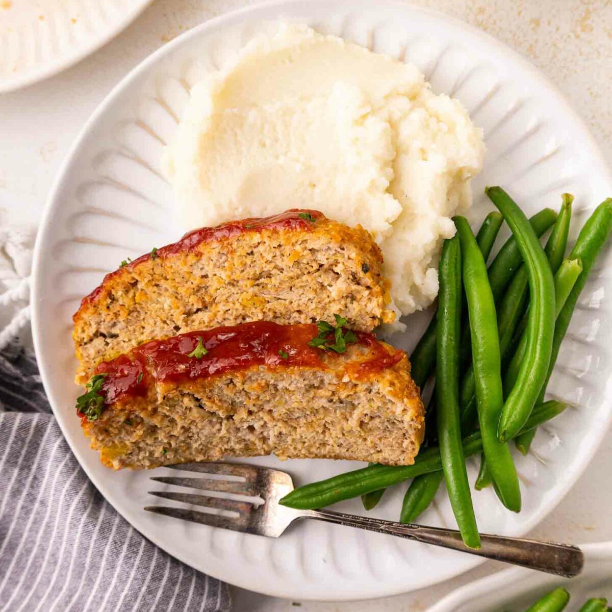 A white plate with two slices of meatloaf topped with ketchup, a serving of mashed potatoes, and a portion of green beans. A fork rests on the plate next to the food.