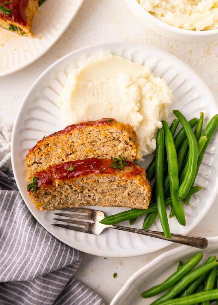 A white plate with two slices of meatloaf topped with ketchup, a serving of mashed potatoes, and a portion of green beans. A fork rests on the plate next to the food.