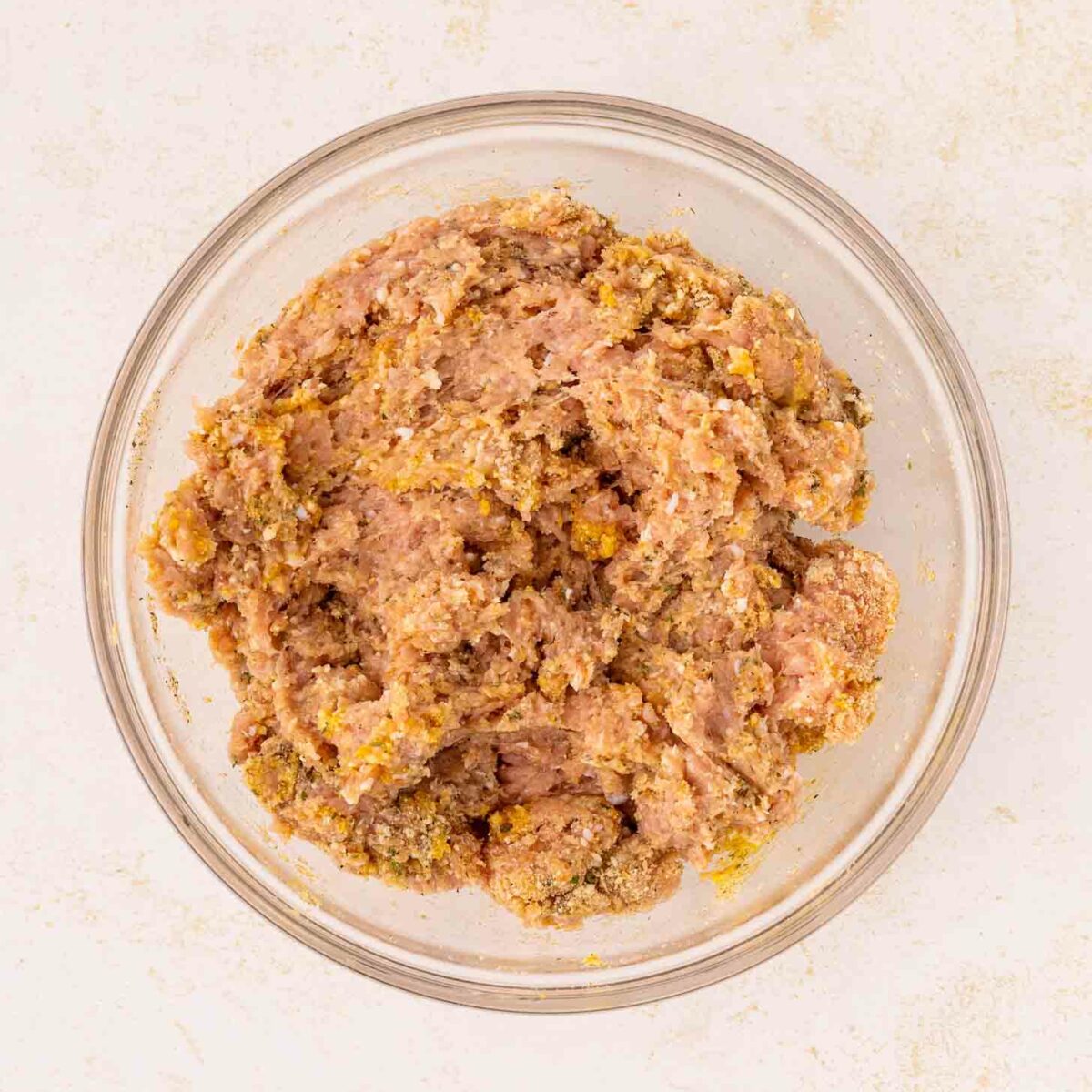 A glass bowl filled with a mixture of raw ground meat and seasonings, placed on a light-colored surface.
