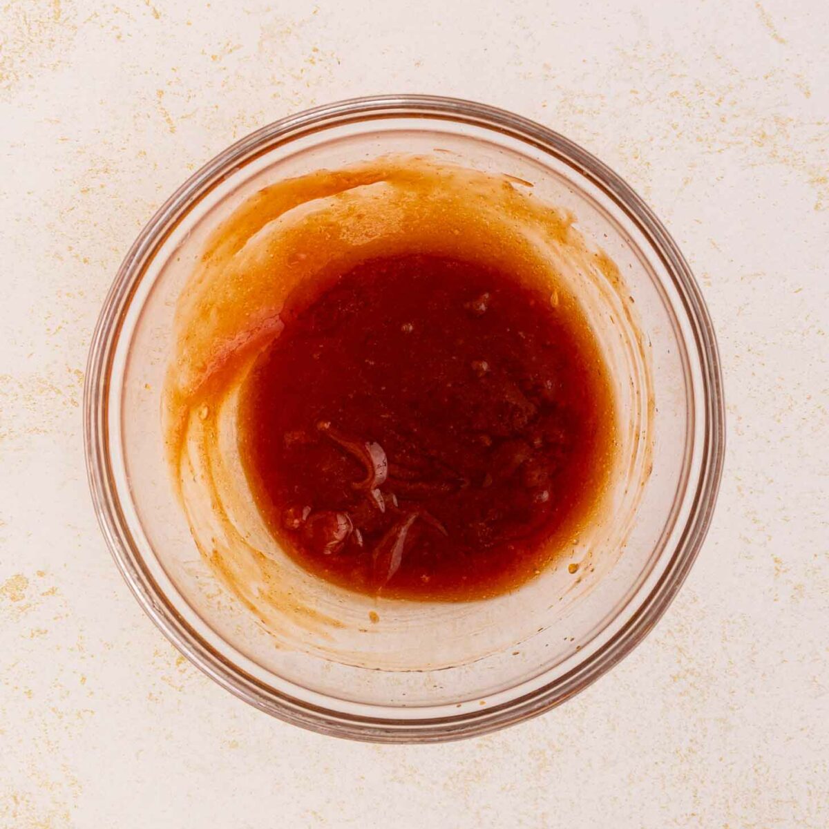 A clear glass bowl filled with a reddish-brown sauce sits on a light, textured surface. Some sauce clings to the sides of the bowl.