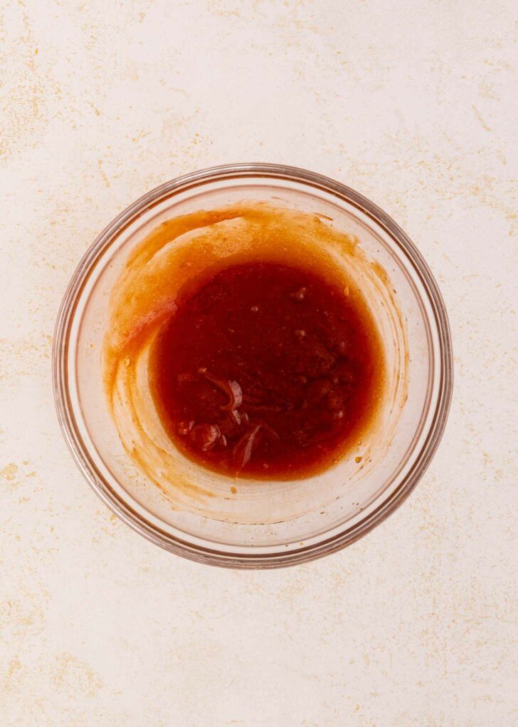 A clear glass bowl filled with a reddish-brown sauce sits on a light, textured surface. Some sauce clings to the sides of the bowl.