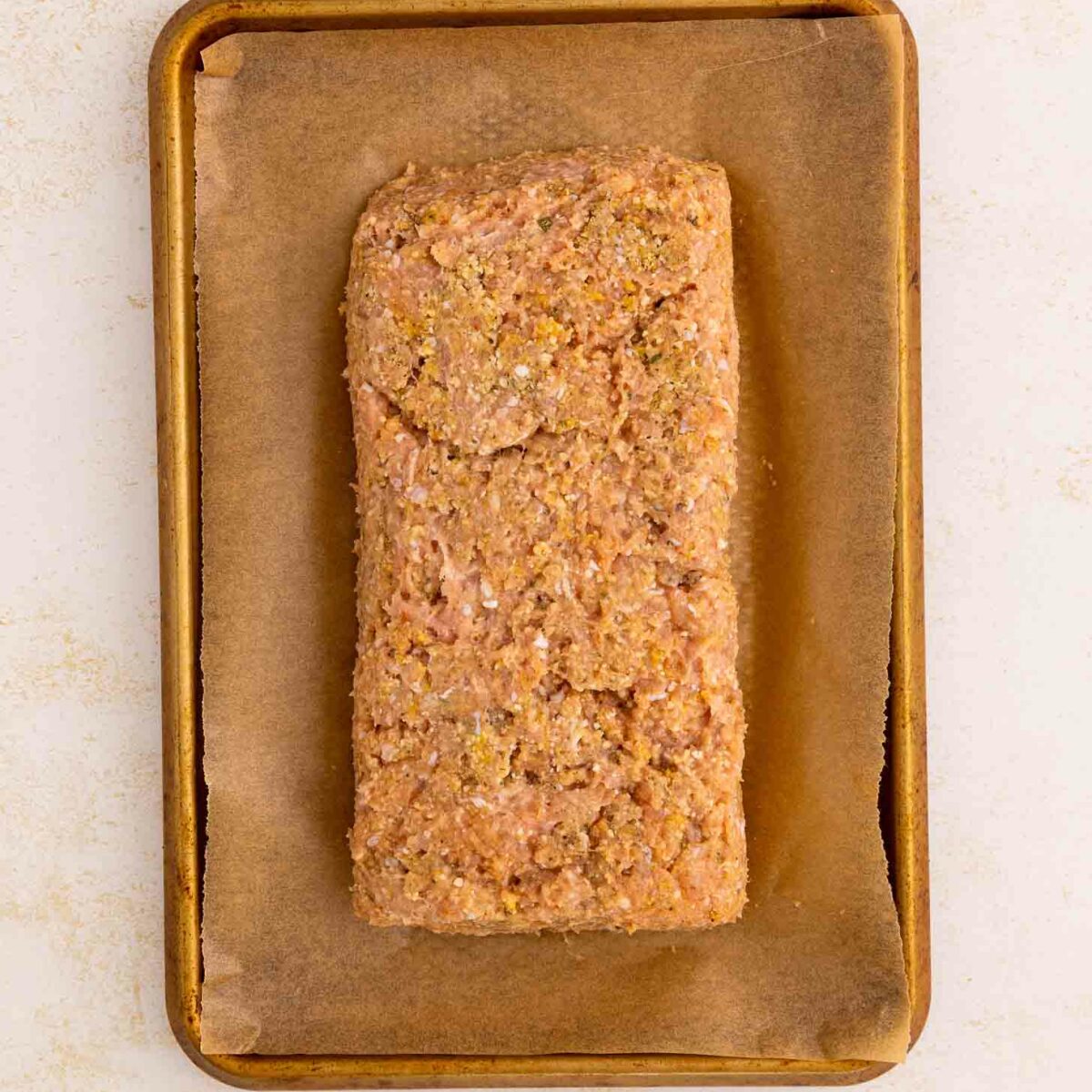 A rectangular, uncooked meatloaf mixture sits on a piece of parchment paper atop a small, rimmed baking sheet against a light background.