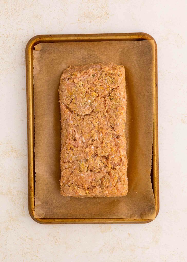 A rectangular, uncooked meatloaf mixture sits on a piece of parchment paper atop a small, rimmed baking sheet against a light background.