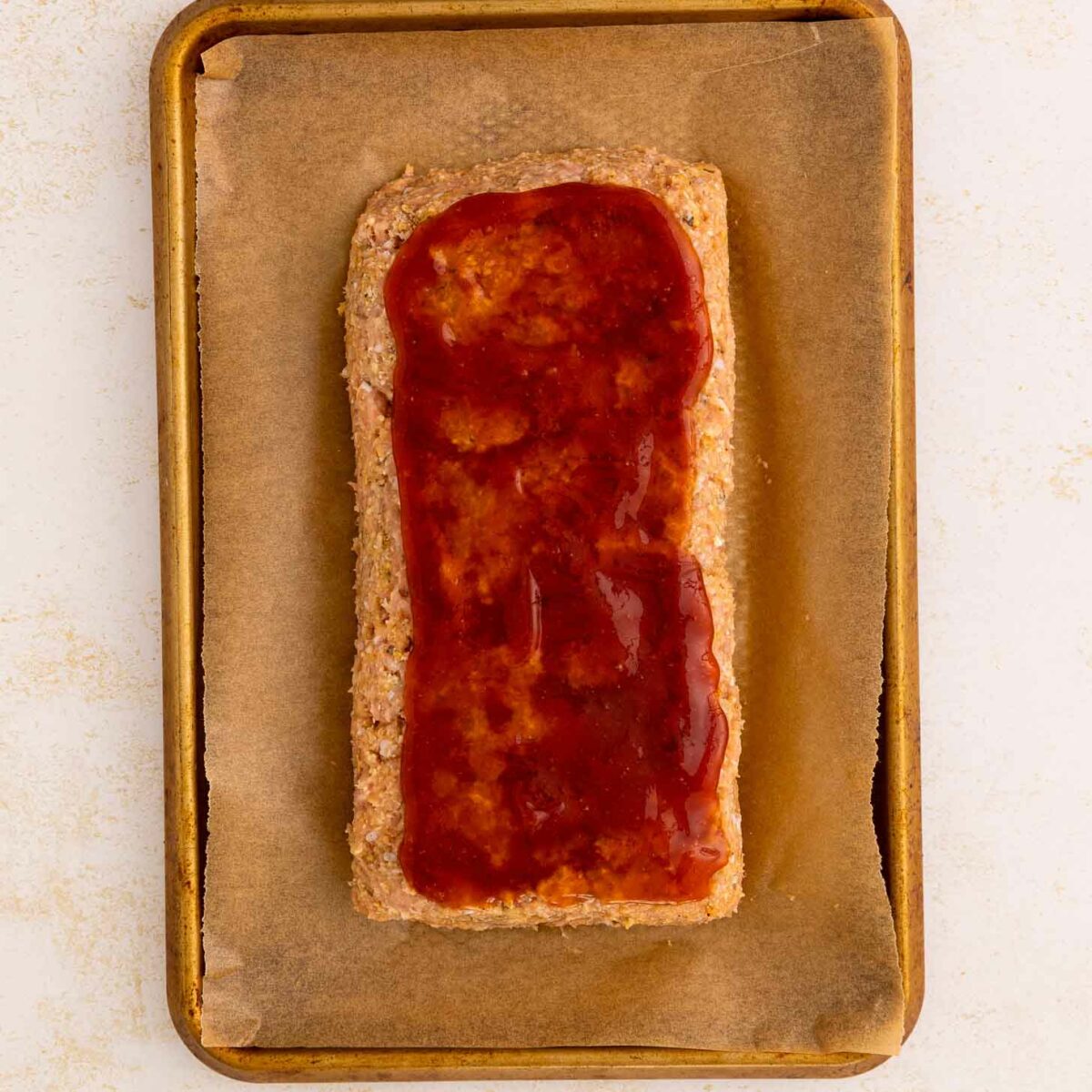 A rectangular meatloaf on a parchment-lined baking sheet, topped with a glossy layer of ketchup and brown sugar mixture.