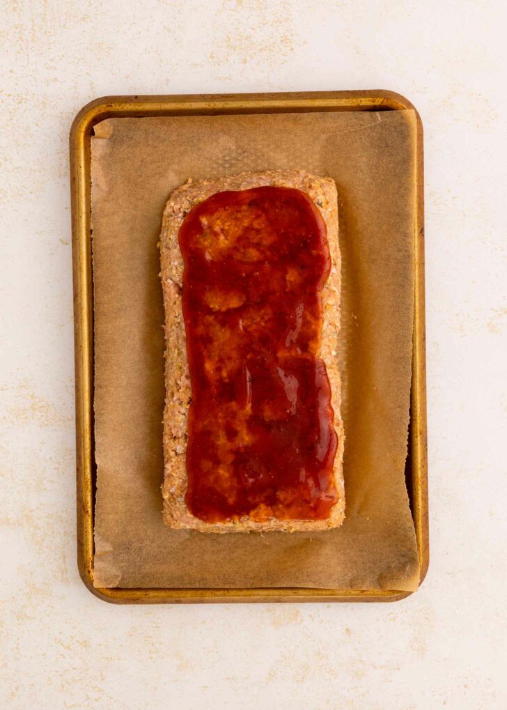 A rectangular meatloaf on a parchment-lined baking sheet, topped with a glossy layer of ketchup and brown sugar mixture.