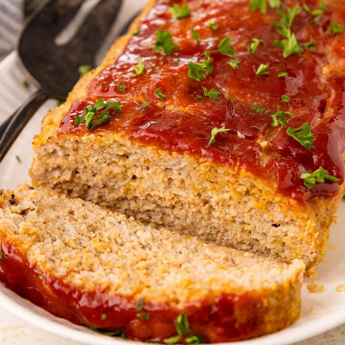 A loaf of chicken meatloaf topped with ketchup glaze and sprinkled with chopped parsley, with one slice cut and served on a white plate.
