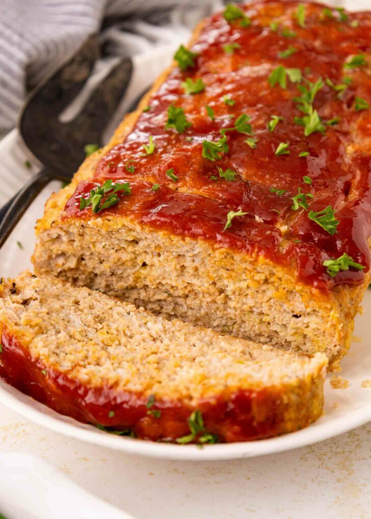 A loaf of chicken meatloaf topped with ketchup glaze and sprinkled with chopped parsley, with one slice cut and served on a white plate.