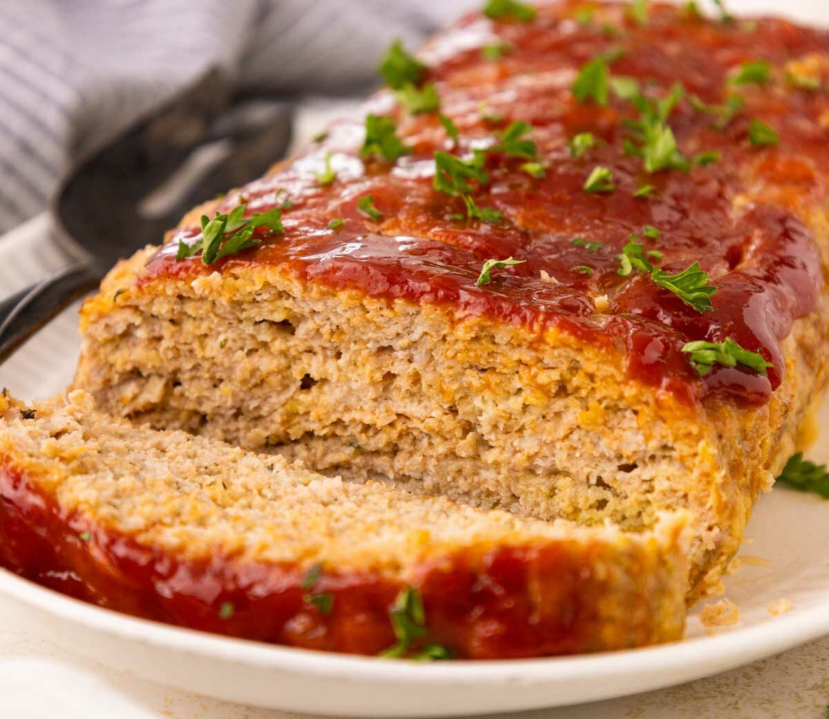 A sliced meatloaf topped with ketchup glaze and garnished with chopped parsley sits on a white plate, with a serving utensil in the background.