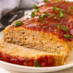 A sliced meatloaf topped with ketchup glaze and garnished with chopped parsley sits on a white plate, with a serving utensil in the background.