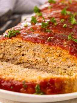 A sliced meatloaf topped with ketchup glaze and garnished with chopped parsley sits on a white plate, with a serving utensil in the background.