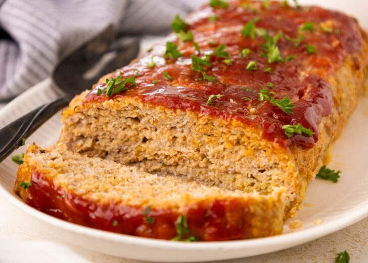 A sliced meatloaf topped with ketchup glaze and garnished with chopped parsley sits on a white plate, with a serving utensil in the background.
