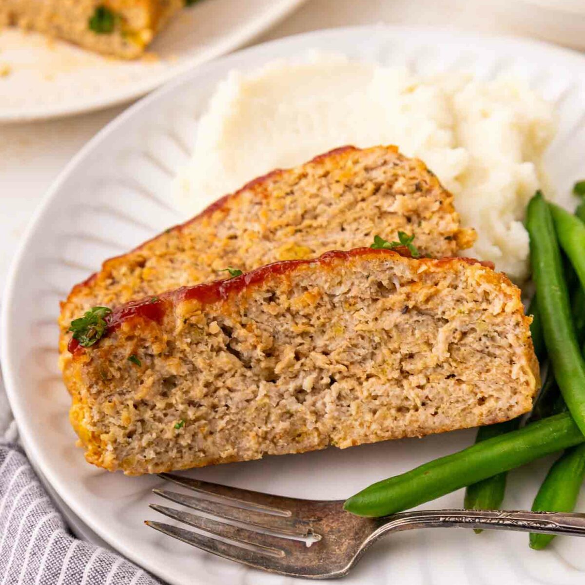 Two slices of meatloaf with a ketchup glaze are served on a white plate with mashed potatoes and green beans. A fork rests on the plate next to a striped napkin.