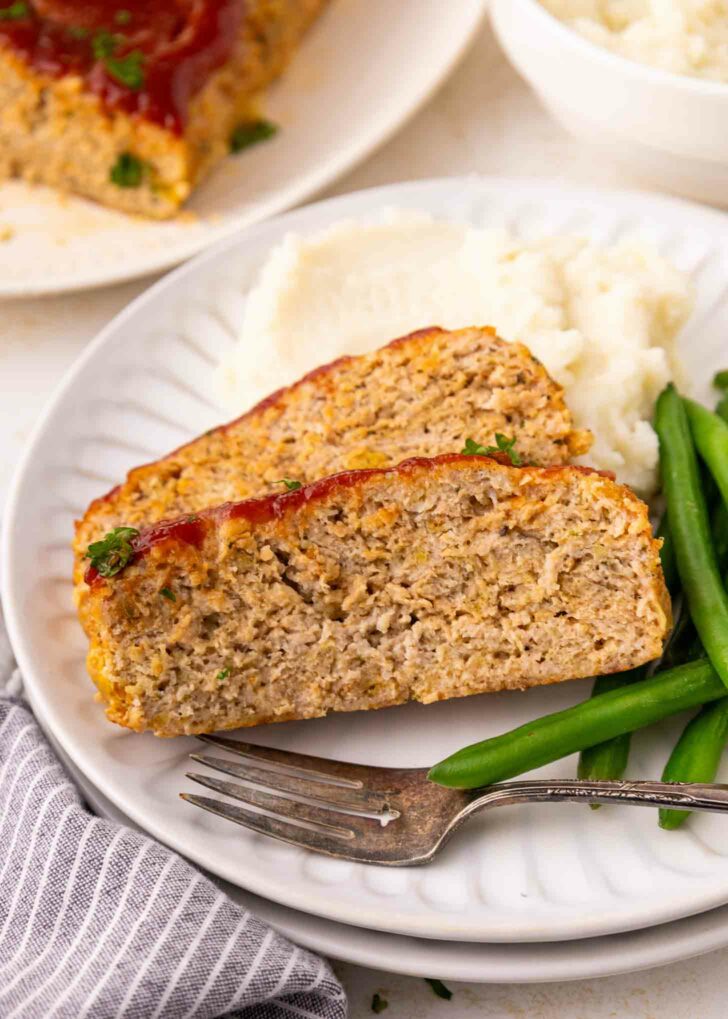 Two slices of meatloaf with a ketchup glaze are served on a white plate with mashed potatoes and green beans. A fork rests on the plate next to a striped napkin.