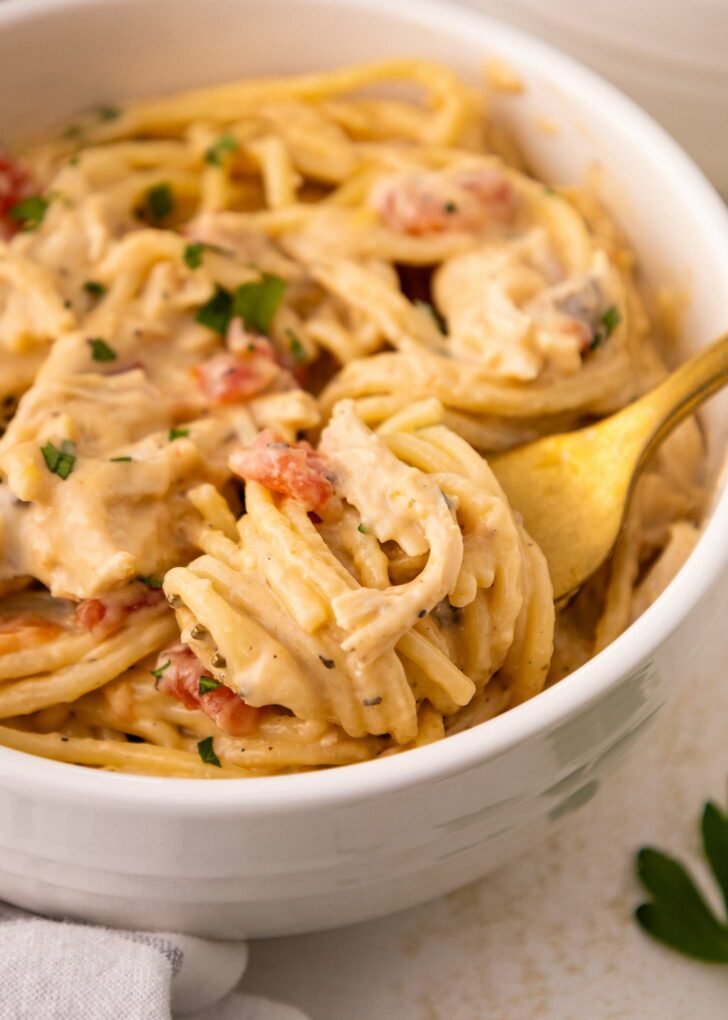 A close-up of creamy spaghetti with pieces of tomato and parsley, served in a white bowl. A golden fork is twirling some of the pasta.
