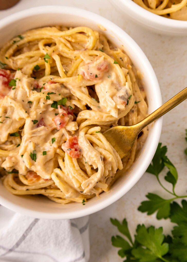 A white bowl filled with creamy spaghetti mixed with shredded chicken, diced tomatoes, and garnished with chopped parsley. A gold fork is twirled into the pasta, and fresh parsley is nearby.