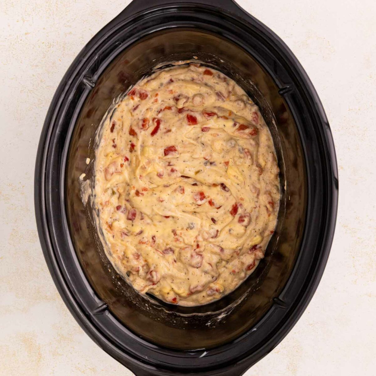 A slow cooker filled with an uncooked mixture of rotel, soup and batter, ready to be cooked. The slow cooker is black and sits on a light-colored surface.