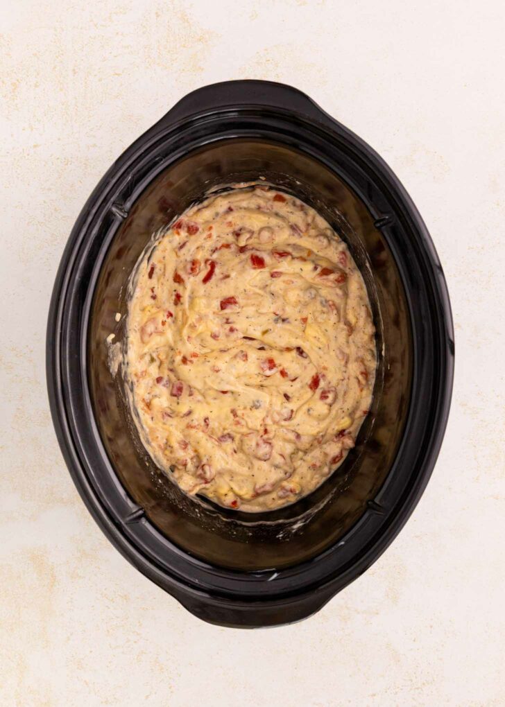 A slow cooker filled with an uncooked mixture of rotel, soup and batter, ready to be cooked. The slow cooker is black and sits on a light-colored surface.
