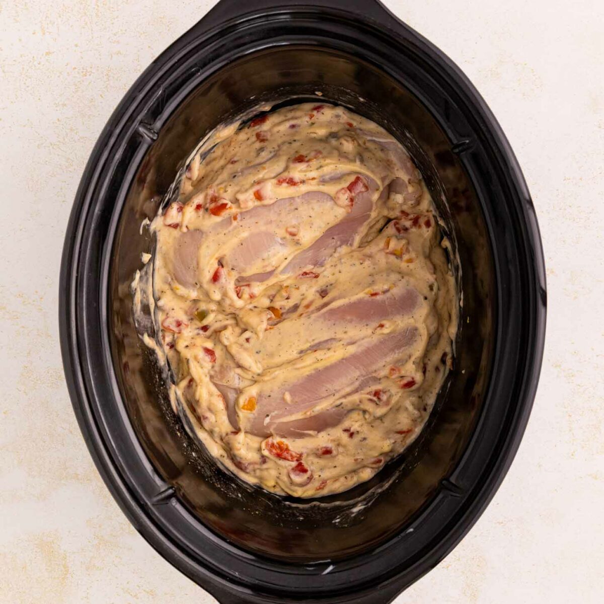 Uncooked chicken breasts covered in a creamy sauce with diced vegetables inside a black slow cooker, viewed from above on a light-colored surface.
