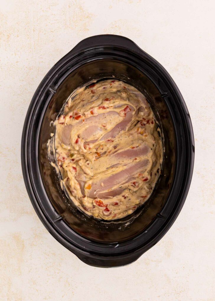 Uncooked chicken breasts covered in a creamy sauce with diced vegetables inside a black slow cooker, viewed from above on a light-colored surface.