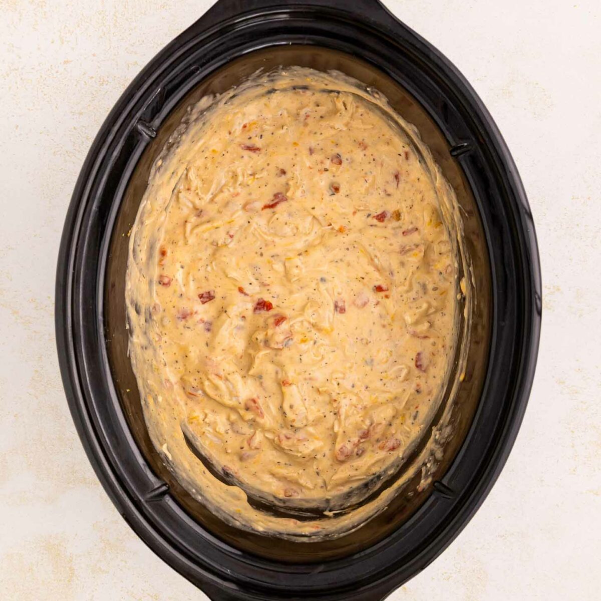 A black slow cooker filled with a creamy, cheesy dip mixture that has visible bits of red and yellow ingredients, sitting on a light-colored surface.