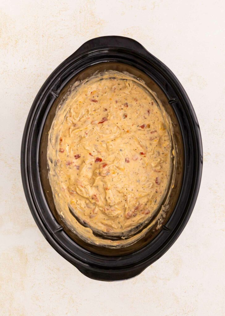 A black slow cooker filled with a creamy, cheesy dip mixture that has visible bits of red and yellow ingredients, sitting on a light-colored surface.