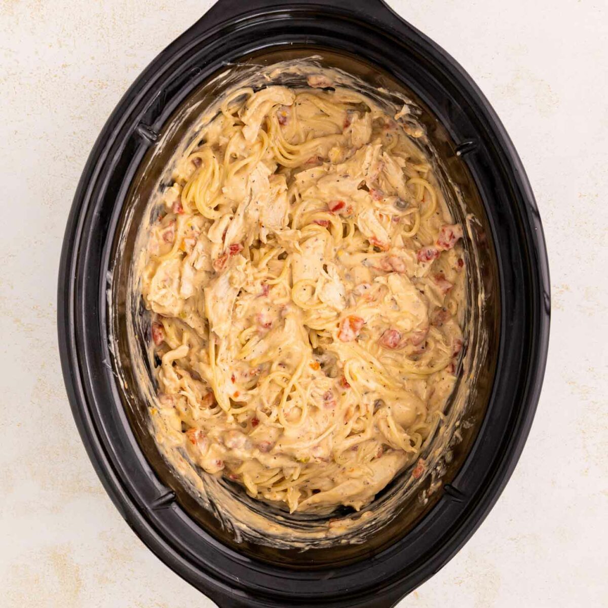 Creamy spaghetti mixed with shredded chicken and diced tomatoes in a black slow cooker, viewed from above on a light-colored surface.