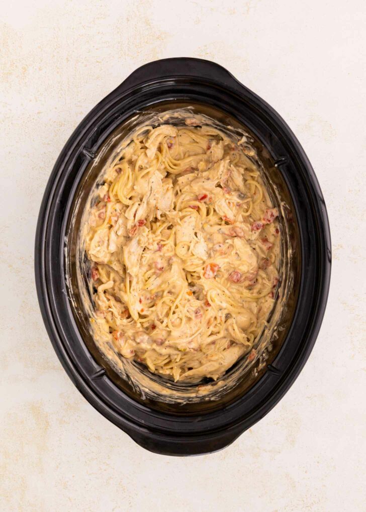Creamy spaghetti mixed with shredded chicken and diced tomatoes in a black slow cooker, viewed from above on a light-colored surface.