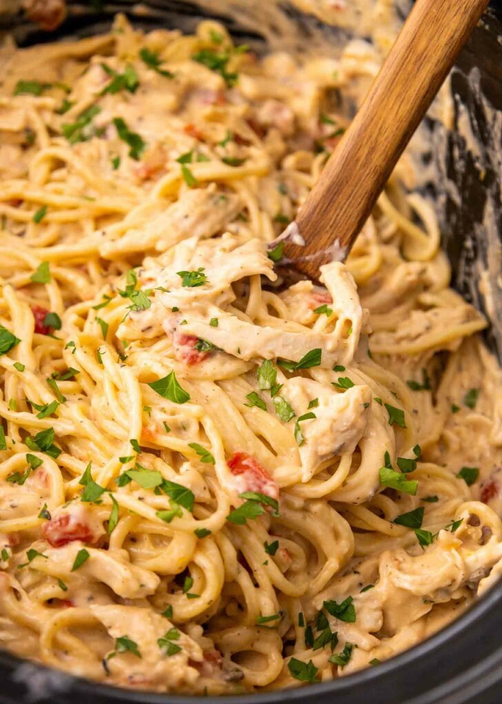 A close-up of creamy pasta with shredded chicken, garnished with chopped parsley, being stirred in a slow cooker with a wooden spoon. The sauce looks rich and includes pieces of tomato.