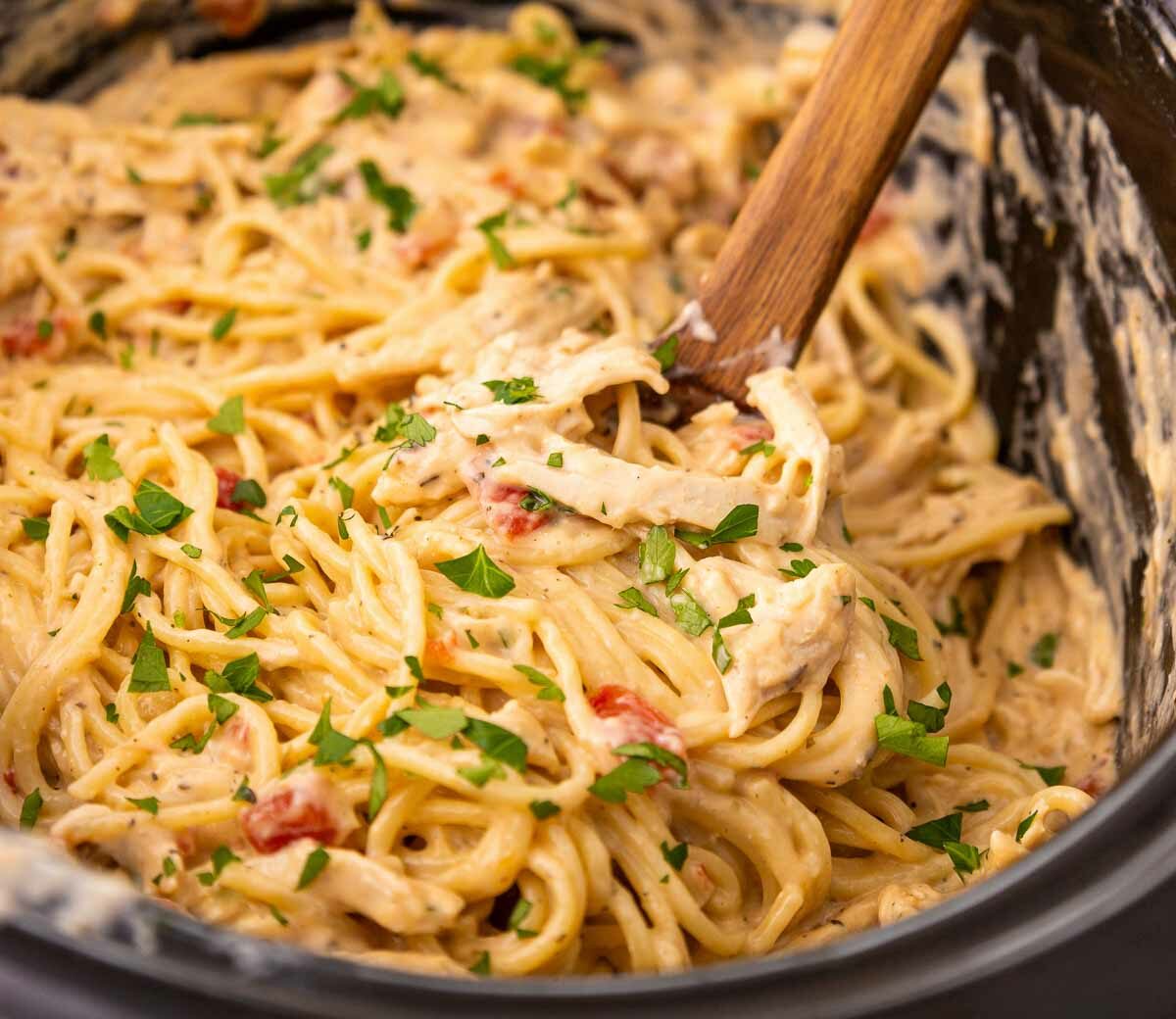 Creamy pasta with shredded chicken, spaghetti noodles, diced tomatoes, and chopped parsley is mixed together in a slow cooker. A wooden spoon is used for stirring the dish.