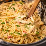 Creamy pasta with shredded chicken, spaghetti noodles, diced tomatoes, and chopped parsley is mixed together in a slow cooker. A wooden spoon is used for stirring the dish.