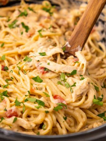Creamy pasta with shredded chicken, spaghetti noodles, diced tomatoes, and chopped parsley is mixed together in a slow cooker. A wooden spoon is used for stirring the dish.