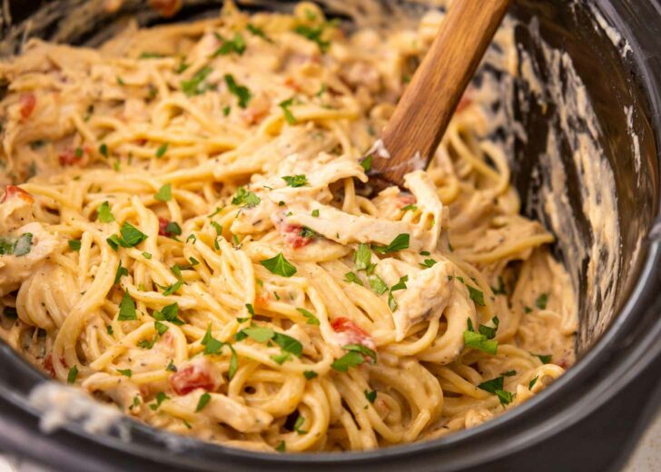 Creamy pasta with shredded chicken, spaghetti noodles, diced tomatoes, and chopped parsley is mixed together in a slow cooker. A wooden spoon is used for stirring the dish.